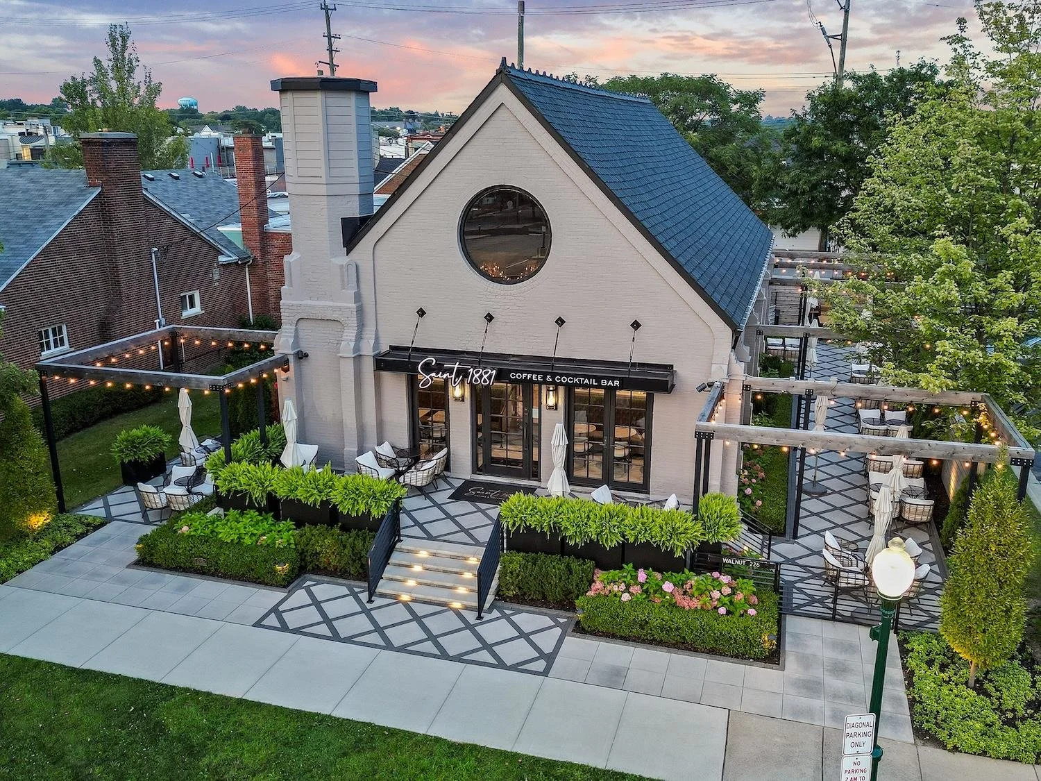 Exterior view of a modern coffee and cocktail bar named 'Saint 1881' with outdoor seating, string lights, and greenery, in an urban area during dusk.