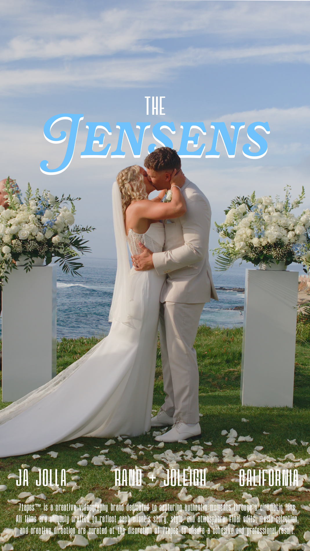 A bride and groom kiss at an outdoor wedding altar overlooking the ocean, with large floral arrangements on either side and white rose petals scattered on the grass.