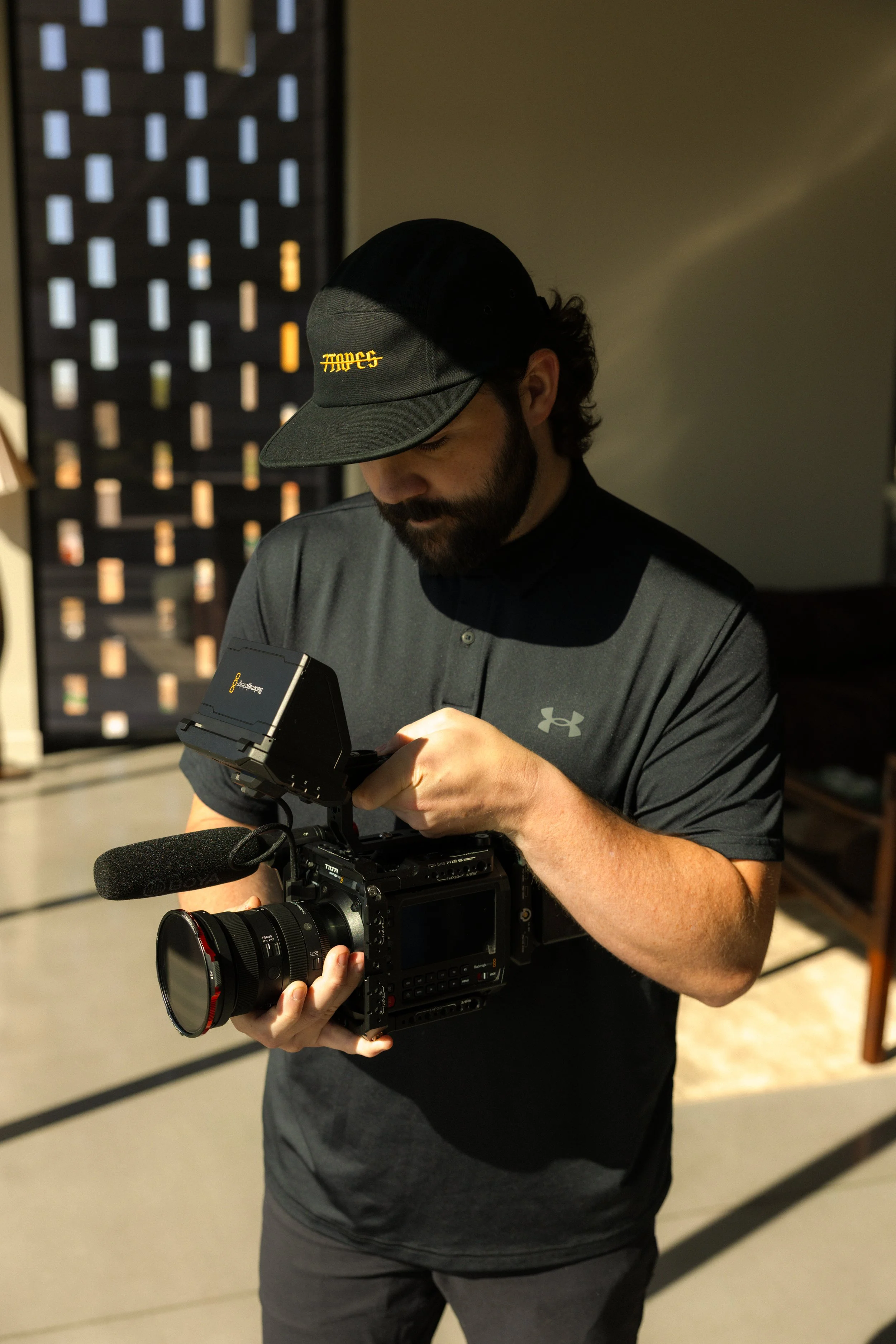 A man with dark hair and a beard wearing a black cap and gray Under Armour shirt is adjusting a professional video camera with a microphone attached, indoors with a decorative wall in the background.