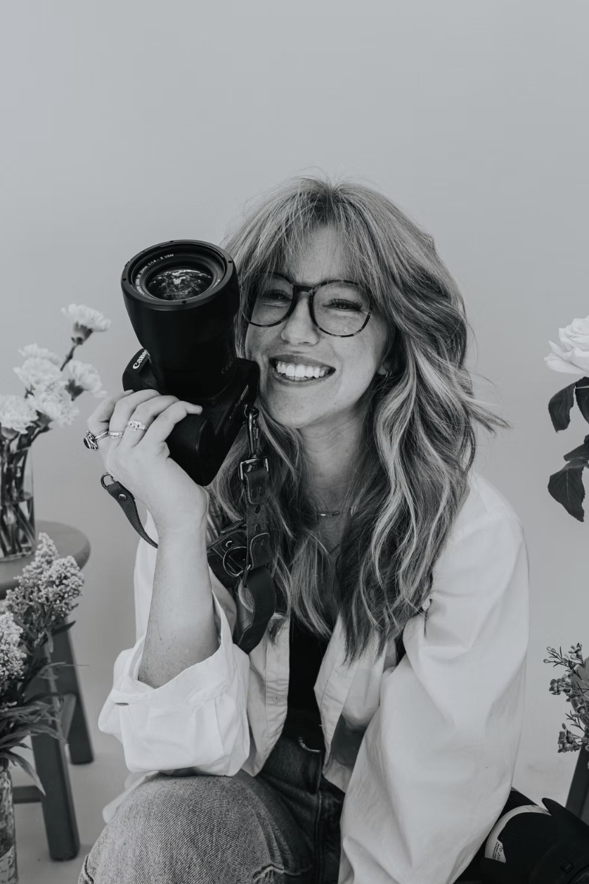 Black and white photo of a woman with wavy hair and glasses smiling while holding a camera up to her eye, surrounded by flowers.