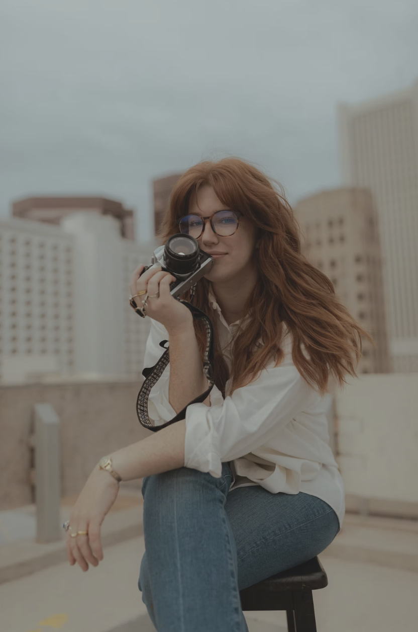 Gillian Rea, photographer. A young woman with long red hair, glasses, and light skin is sitting on a rooftop with city buildings in the background, holding a camera near her face.