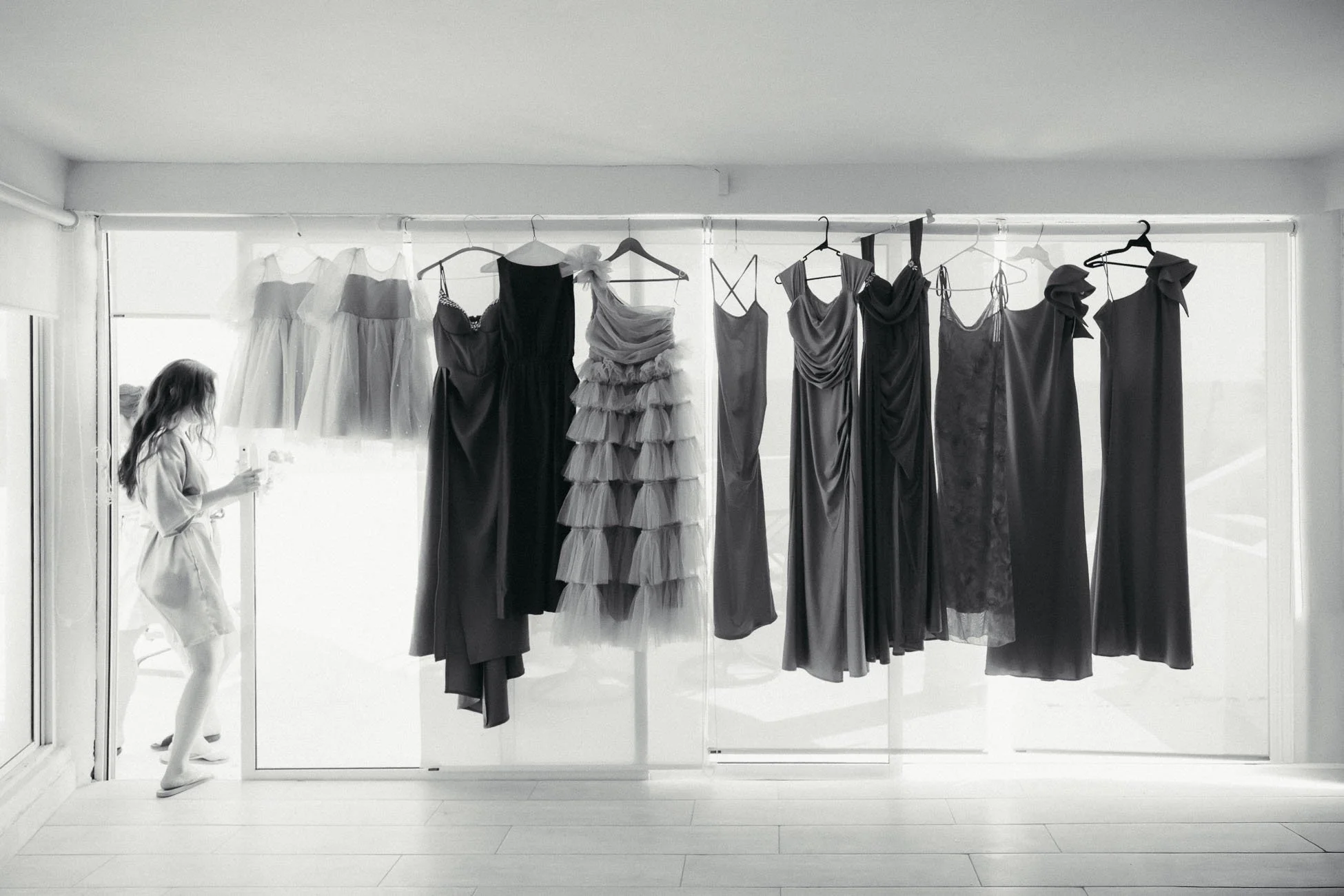 black and white image of dresses hanging on a large glass wall while a woman in a robe walks out