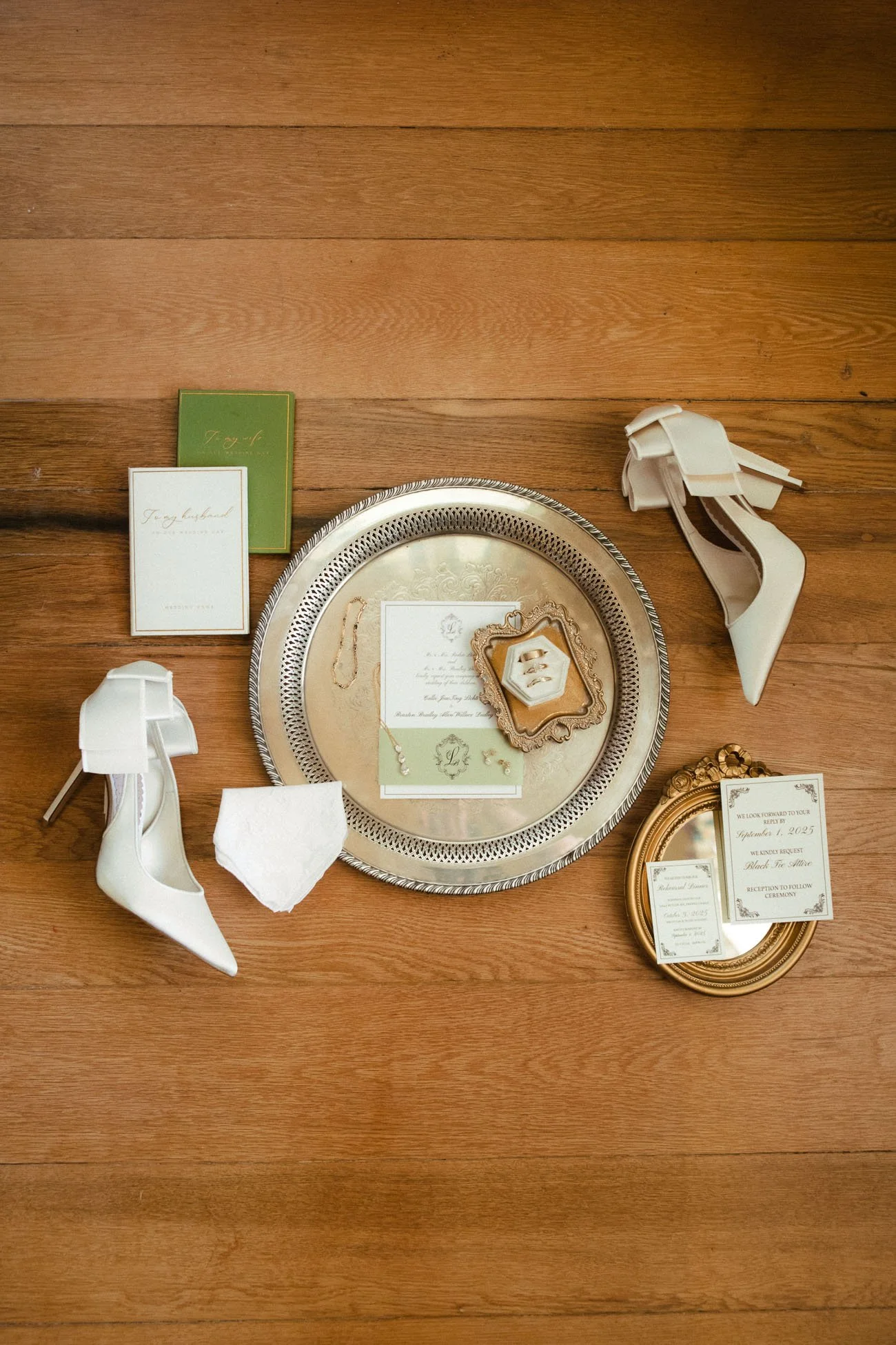 flat lay on wooden floor of silver round tray holding wedding invite and gold dish with rings, surrounded by white heels and wedding vows and sentimental items