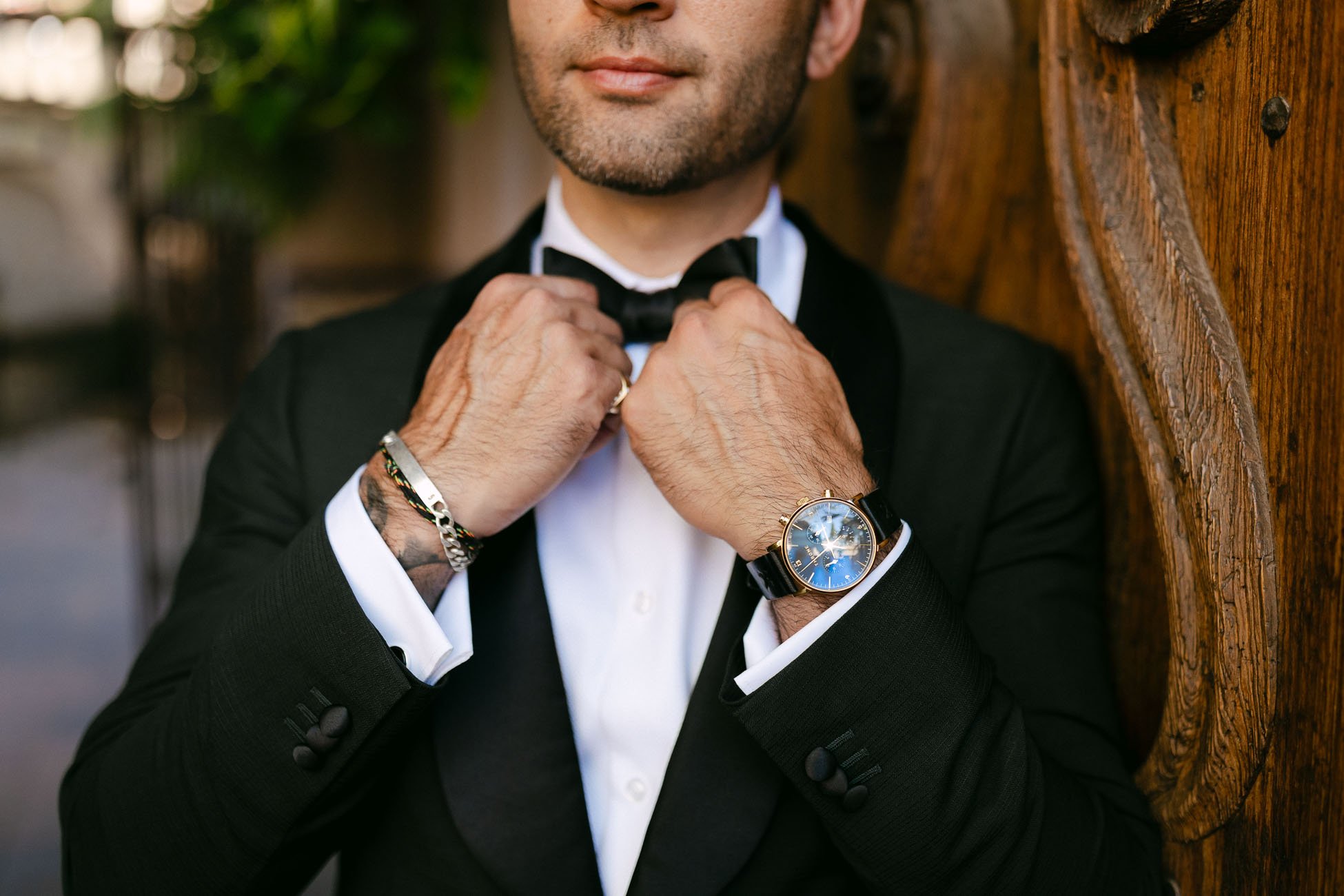 man wearing a black suit and bracelets and a black watch adjusting his bow tie