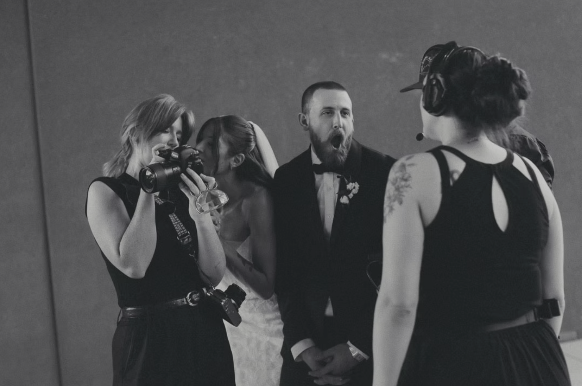 A group of five people at a social event, dressed in formal and casual attire, engaged in conversation against a plain background. One woman is holding a camera, and another woman is whispering to a man in a tuxedo. The focus is on their interactions.