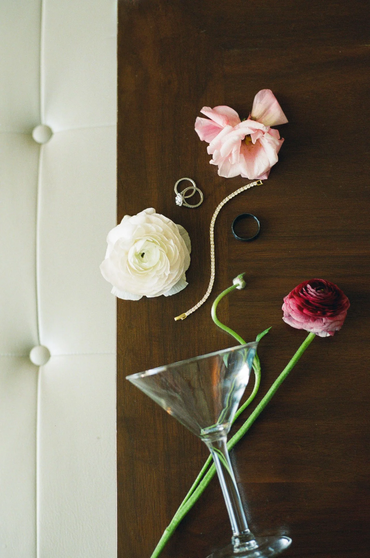 flat lay of flowers, engagement diamond ring, black wedding band, diamond tennis bracelet, roses, and martini glass laying on their side on table