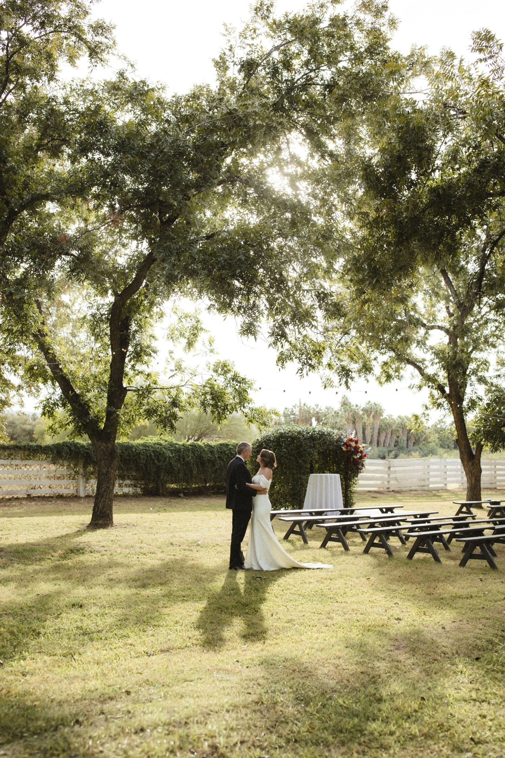 man in suit and woman in white wedding dress embrace in a lush green lawn with huge trees and a white fence