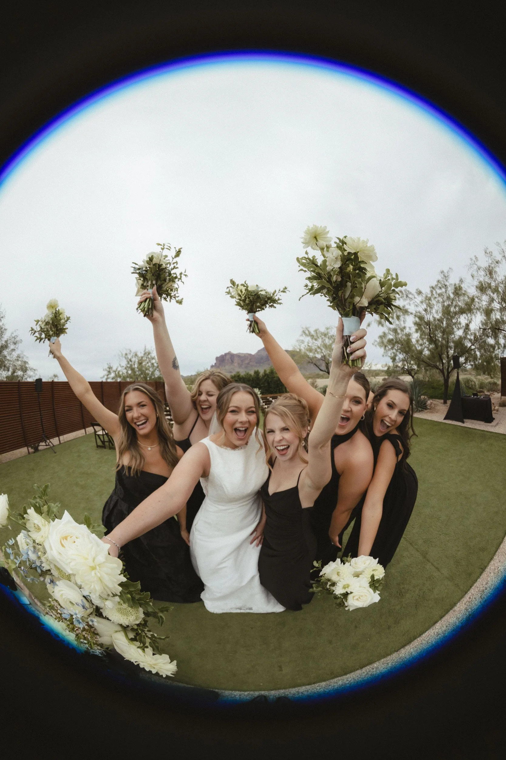 bride and her bridesmaids posing for a fish eye lens camera with their arms stretched out holding white flowers