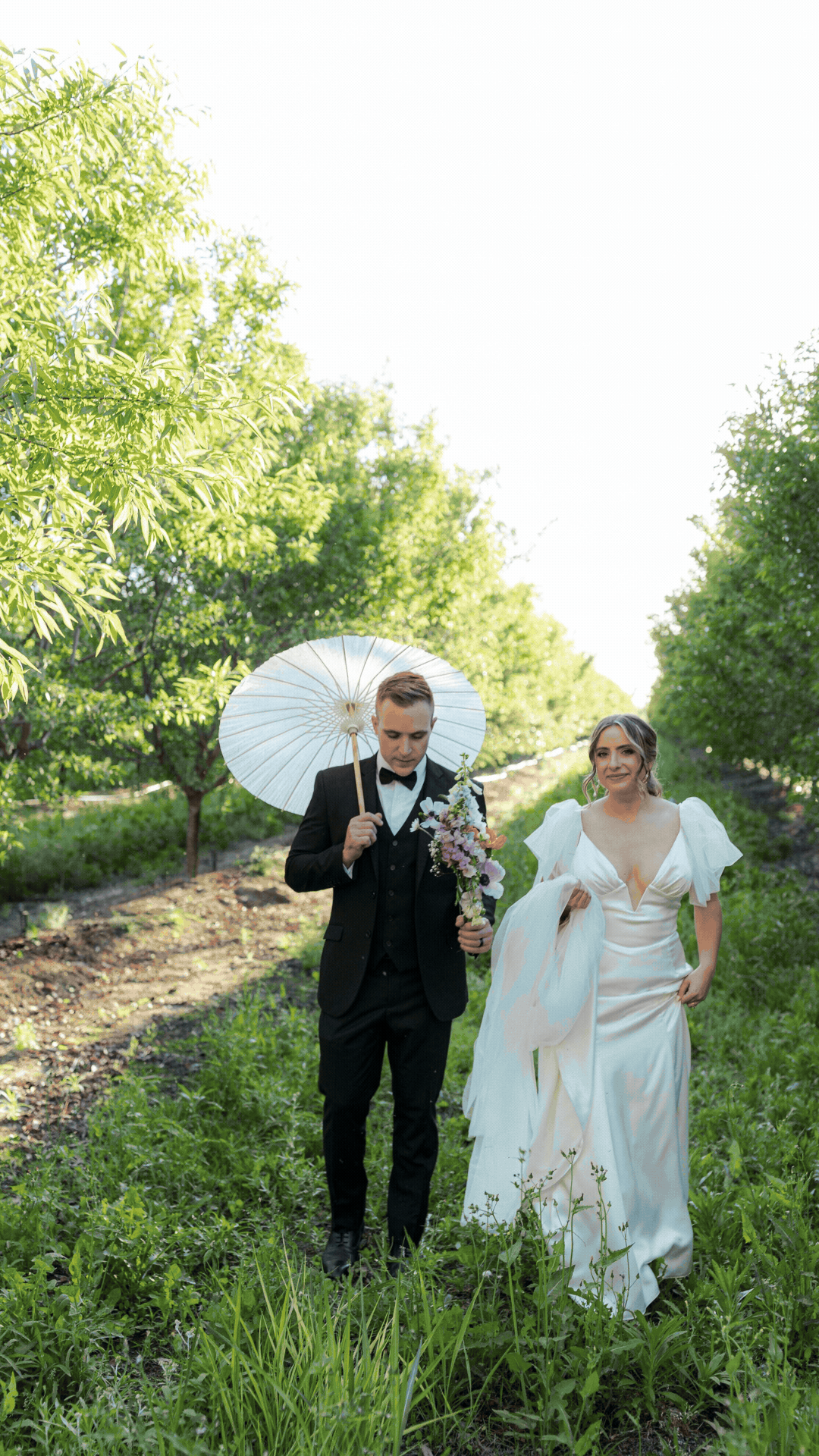 A bride and groom walking through a green outdoor area, with the groom holding a white parasol and a bouquet, during daytime.