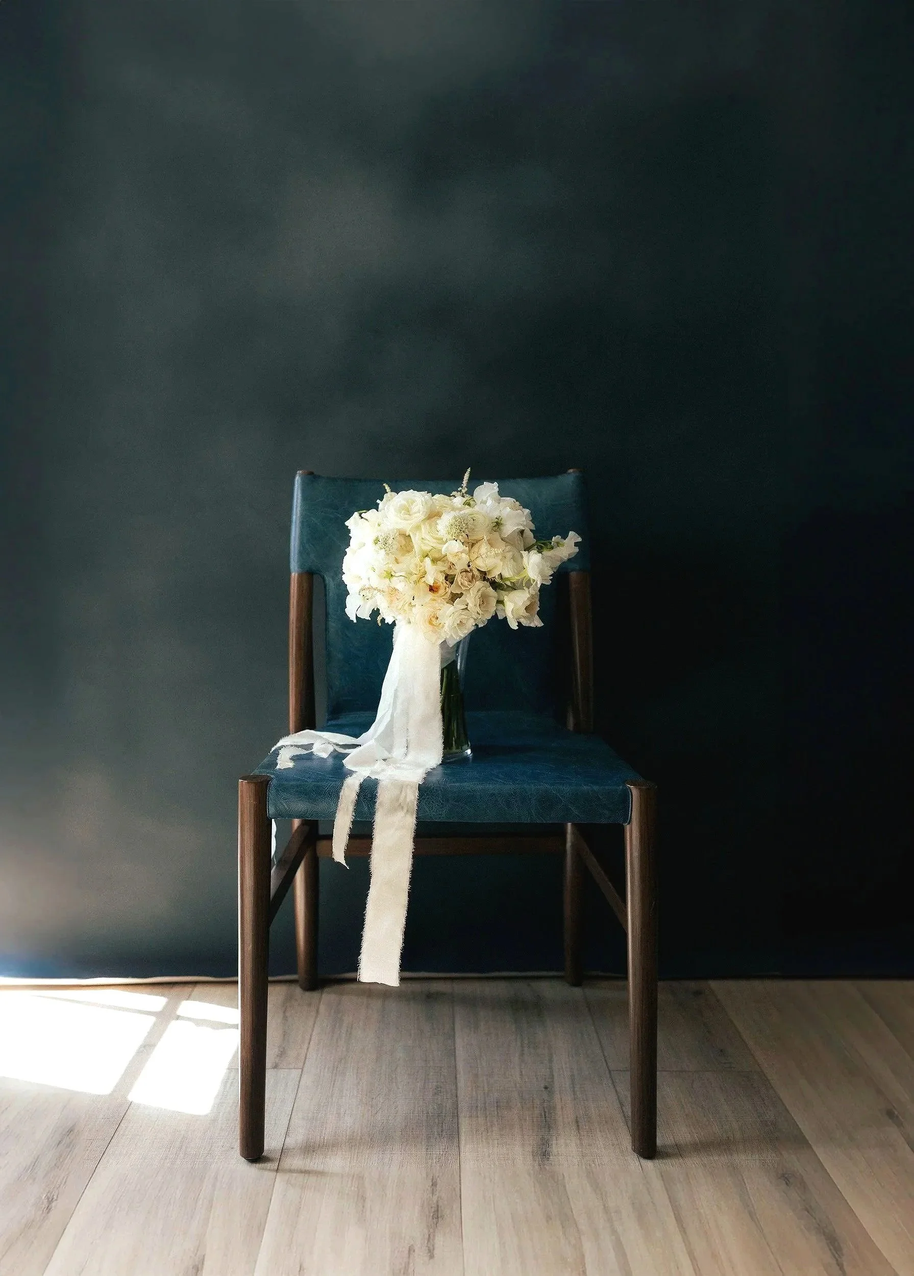 blue leather chair with white bouquet wrapped in white ribbon sitting on it next to a window light