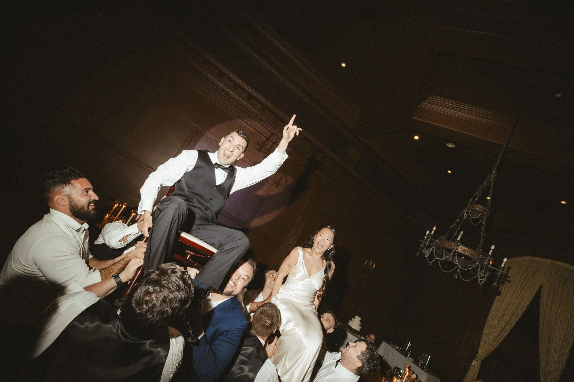 man in black vest pointing to the ceiling while being lifted on a chair in the middle of dance circle