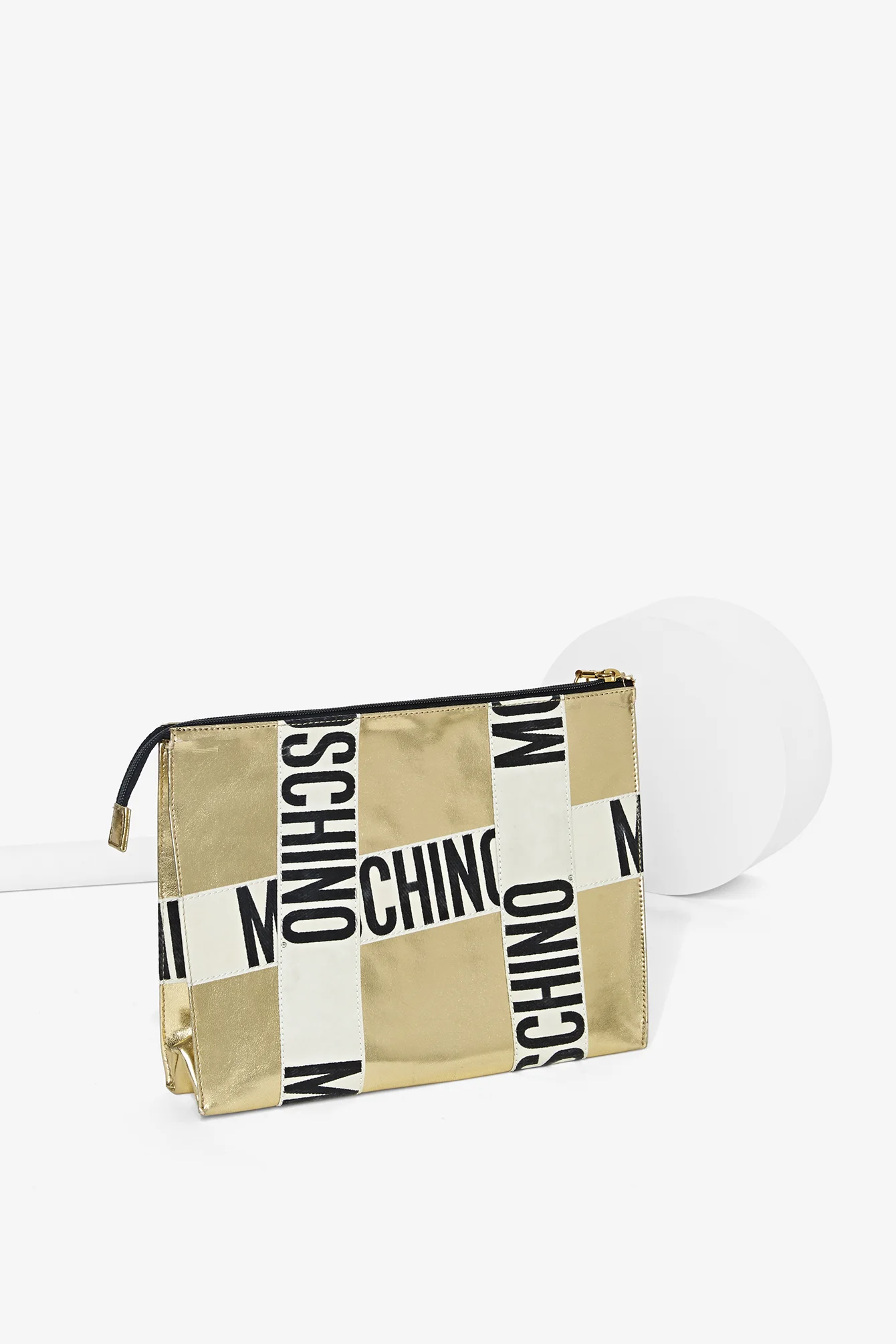 A gold-colored pouch with black, white, and gold striped straps featuring the word 'MOSCHINO' printed repeatedly in bold black letters, positioned on a white background with a white spherical object beside it.