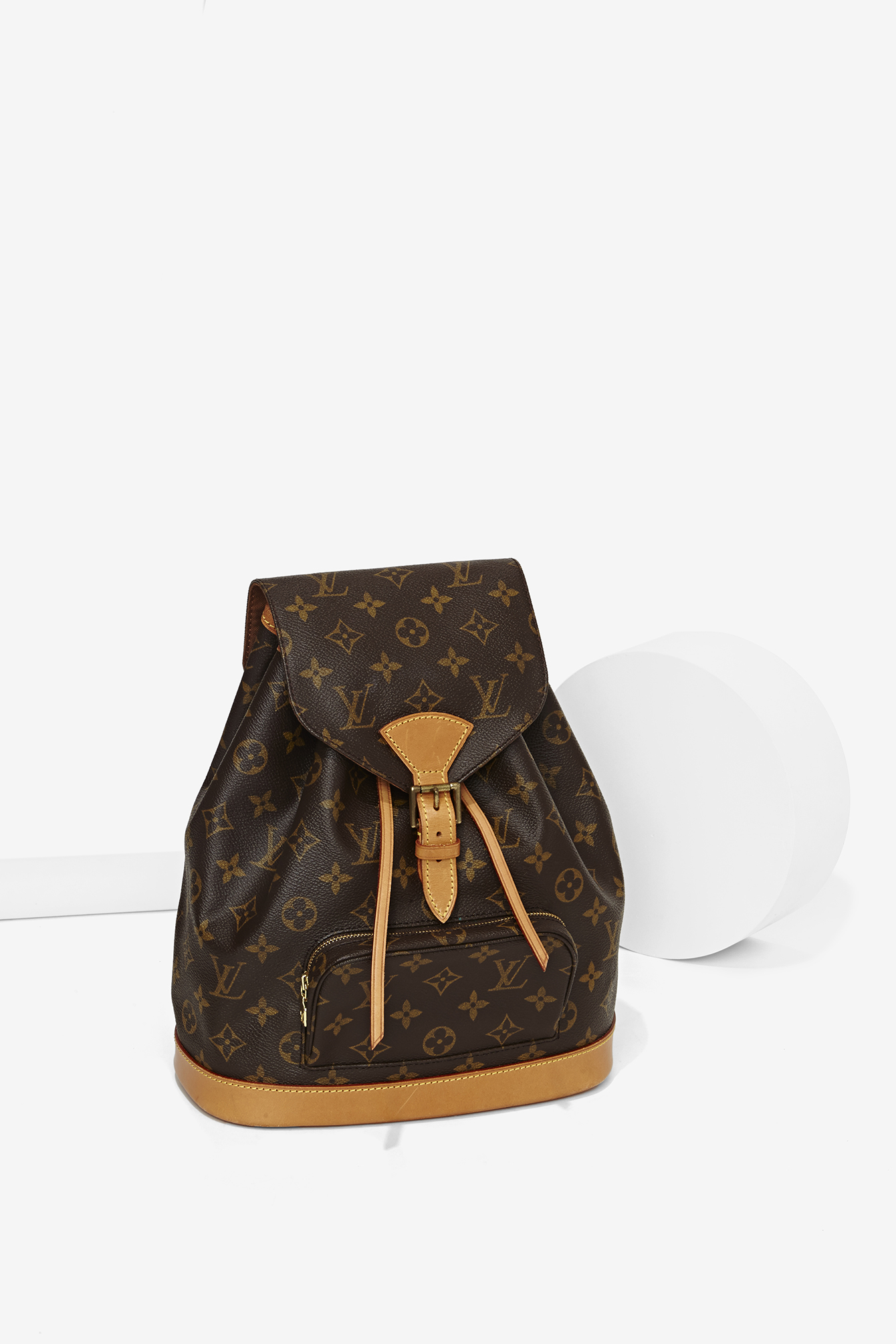 A Louis Vuitton monogram canvas backpack with tan leather accents, placed on a white surface with abstract white shapes in the background. e-commerce, editorial, fashion, vogue