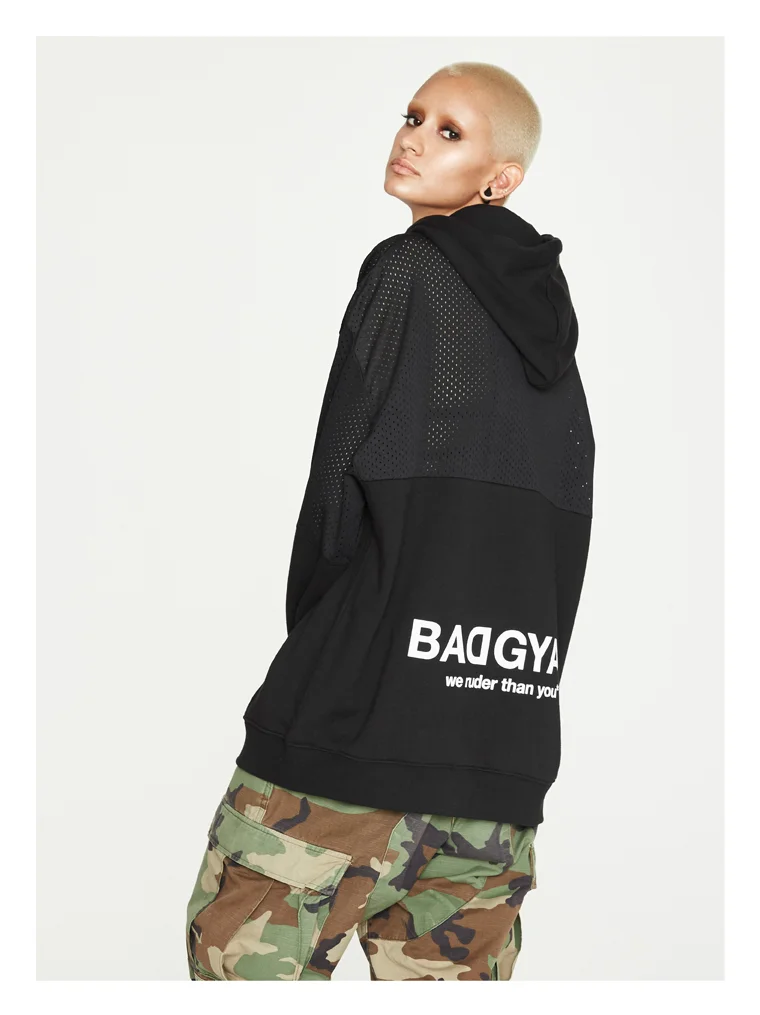 Person with bleached blonde hair and earrings wearing a black hoodie with mesh details and lettering, camo pants, standing against a plain background.