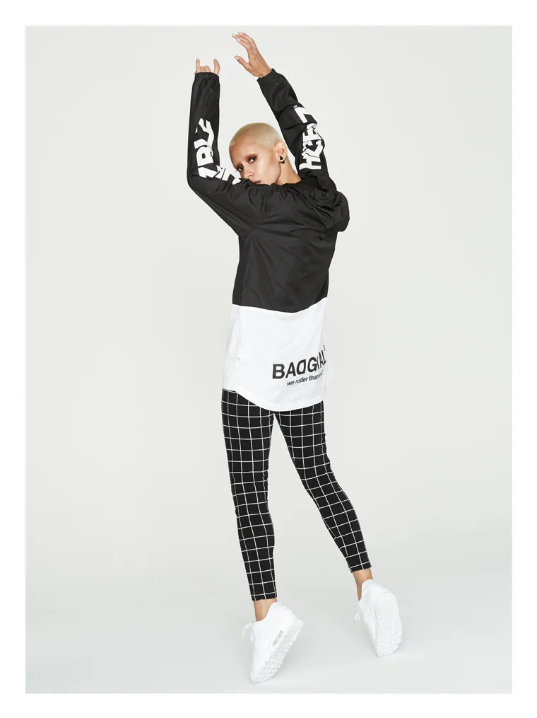 A person with a shaved head wearing a black and white color-blocked jacket, black and white checkered leggings, and white sneakers, posing with arms raised in a studio setting.