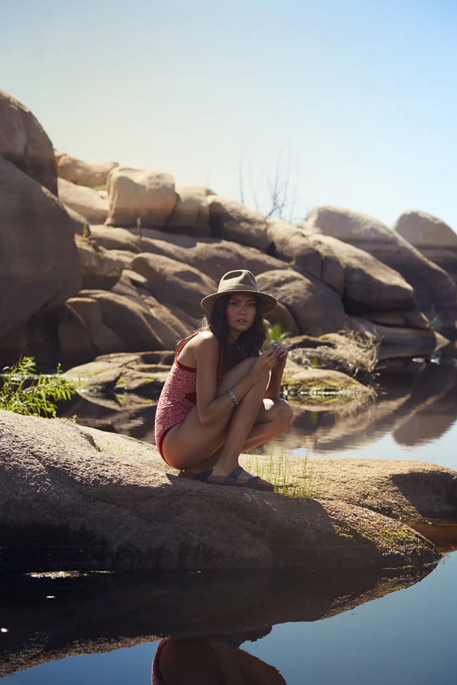 A young woman in a red swimsuit and a wide-brimmed hat crouches on a large rock by a calm river, surrounded by rocks and sparse vegetation, with clear water reflecting her and the sky.