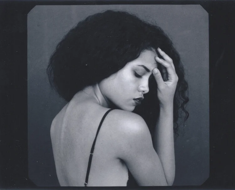 A black and white photograph of a woman with curly hair, her eyes closed, and her hand resting on her forehead in a contemplative pose.