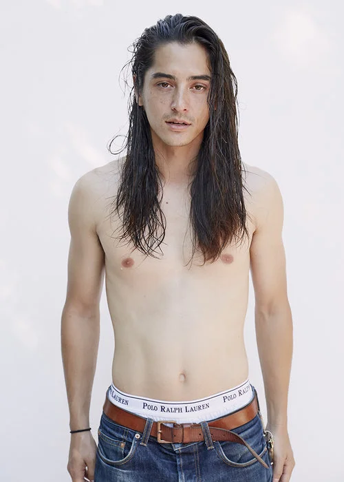 A young man with long dark hair, topless, wearing blue jeans and a brown belt, standing against a plain white background.