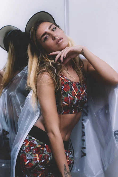 Young woman with long wavy hair, wearing a colorful patterned crop top and matching shorts, standing against a mirror with a white wall in the background, posing with her hand near her face.