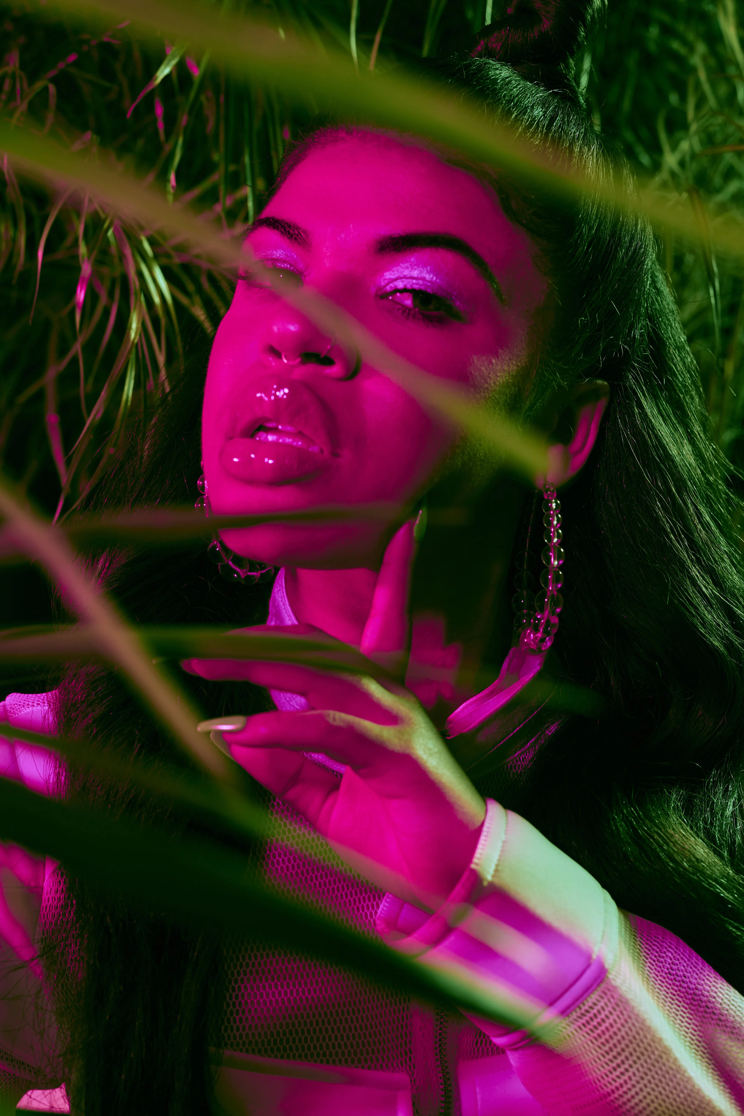 A woman with dark hair and makeup posing among green tropical plants, illuminated with pink and green lighting.