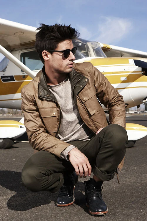 A young man with dark hair, wearing sunglasses, a brown jacket, and dark pants, squatting in front of a small yellow and white aircraft on an airport tarmac.