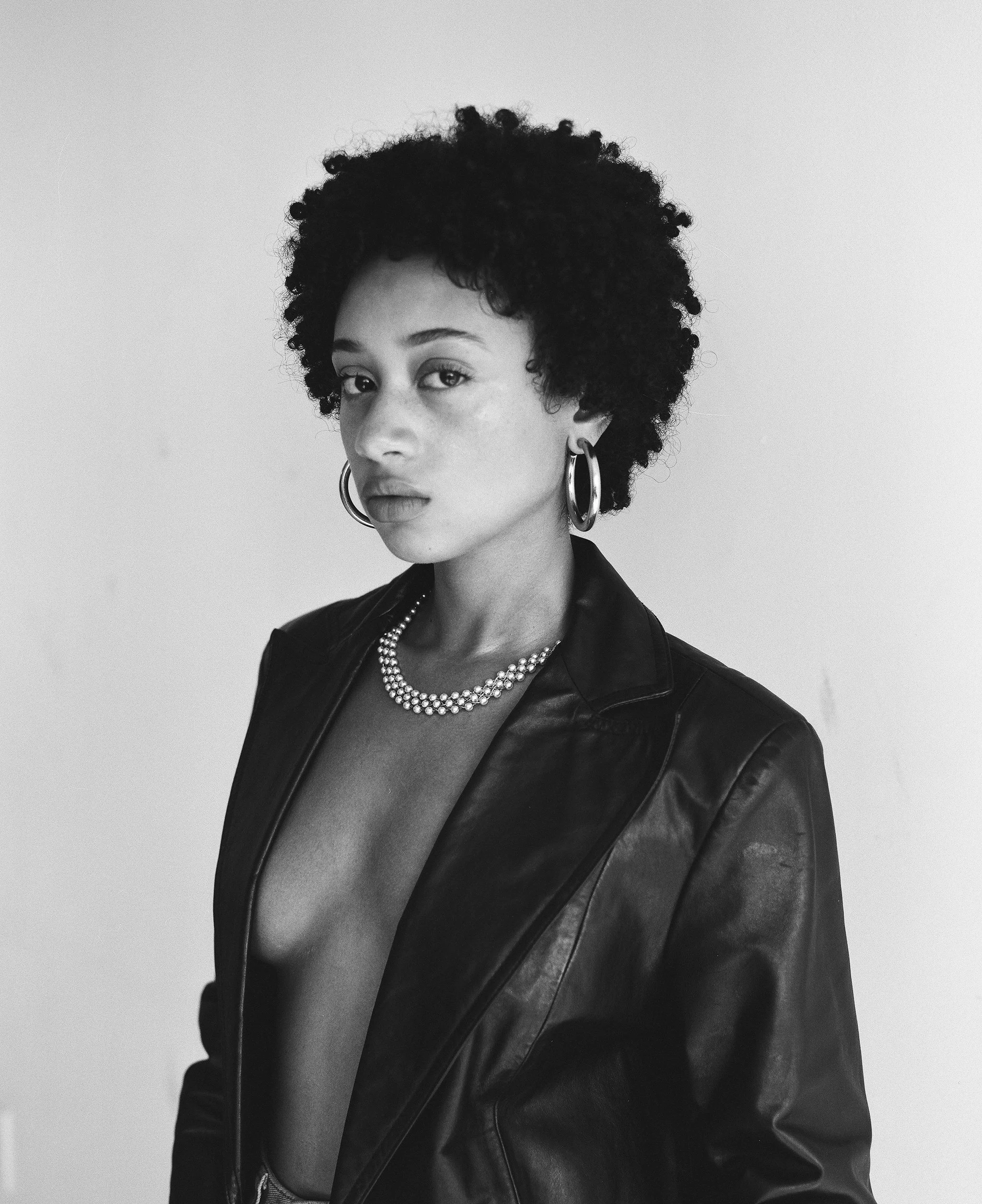 Black and white portrait of a woman with natural curly hair, wearing hoop earrings, a pearl necklace, and a leather jacket, against a plain background.