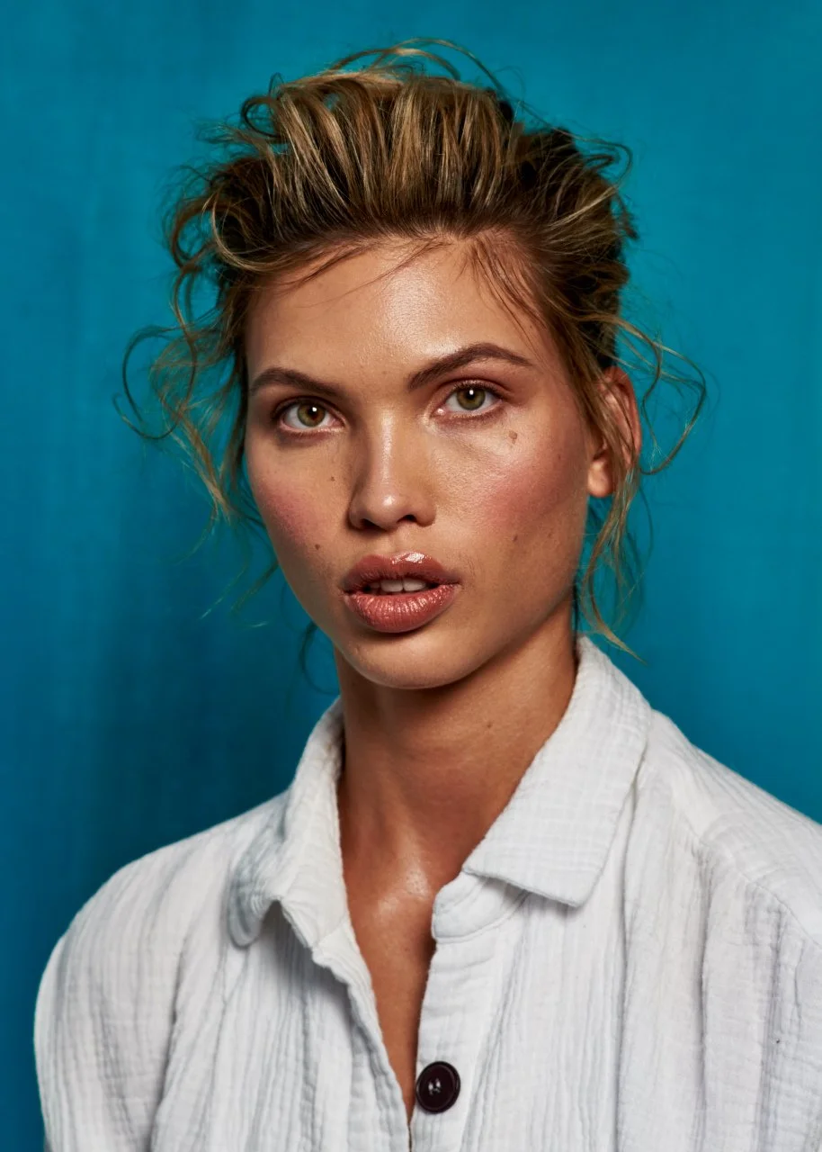 Close-up of a woman with tousled blonde hair, natural makeup, and a white shirt with a button, standing against a blue background.