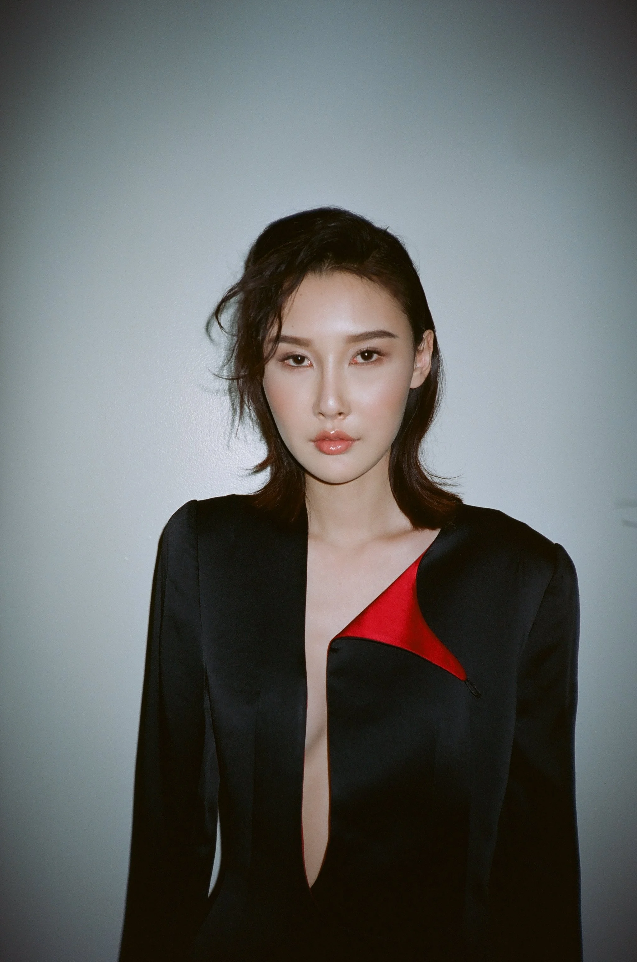A woman with shoulder-length dark hair and fair skin stands against a plain light-colored background. She is wearing a black blazer with a red lapel detail and has a confident expression.