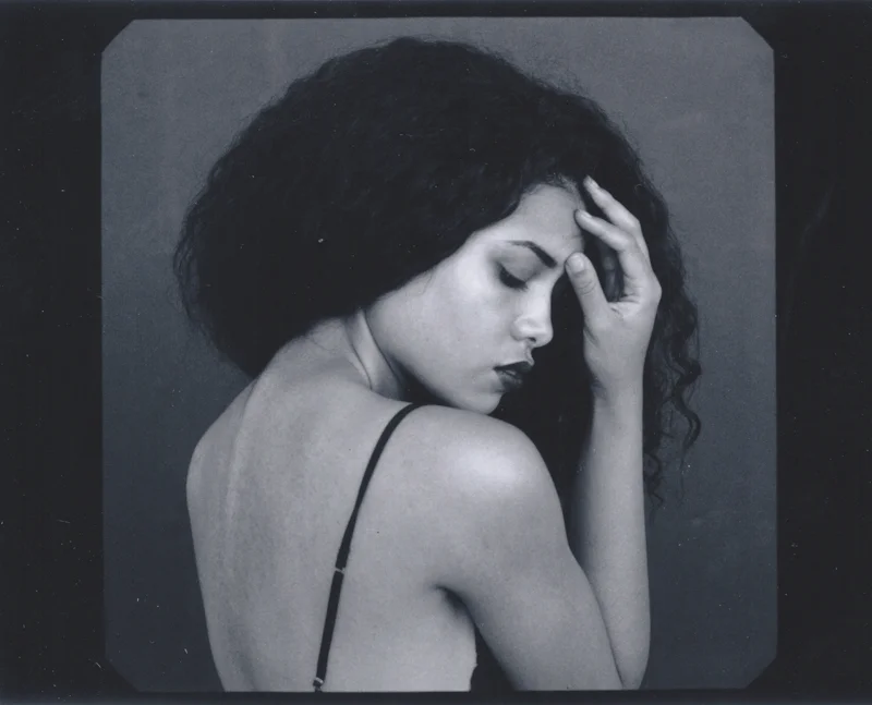 Black and white portrait of a woman with curly hair, eyes closed, hand on her face, wearing a spaghetti strap top.