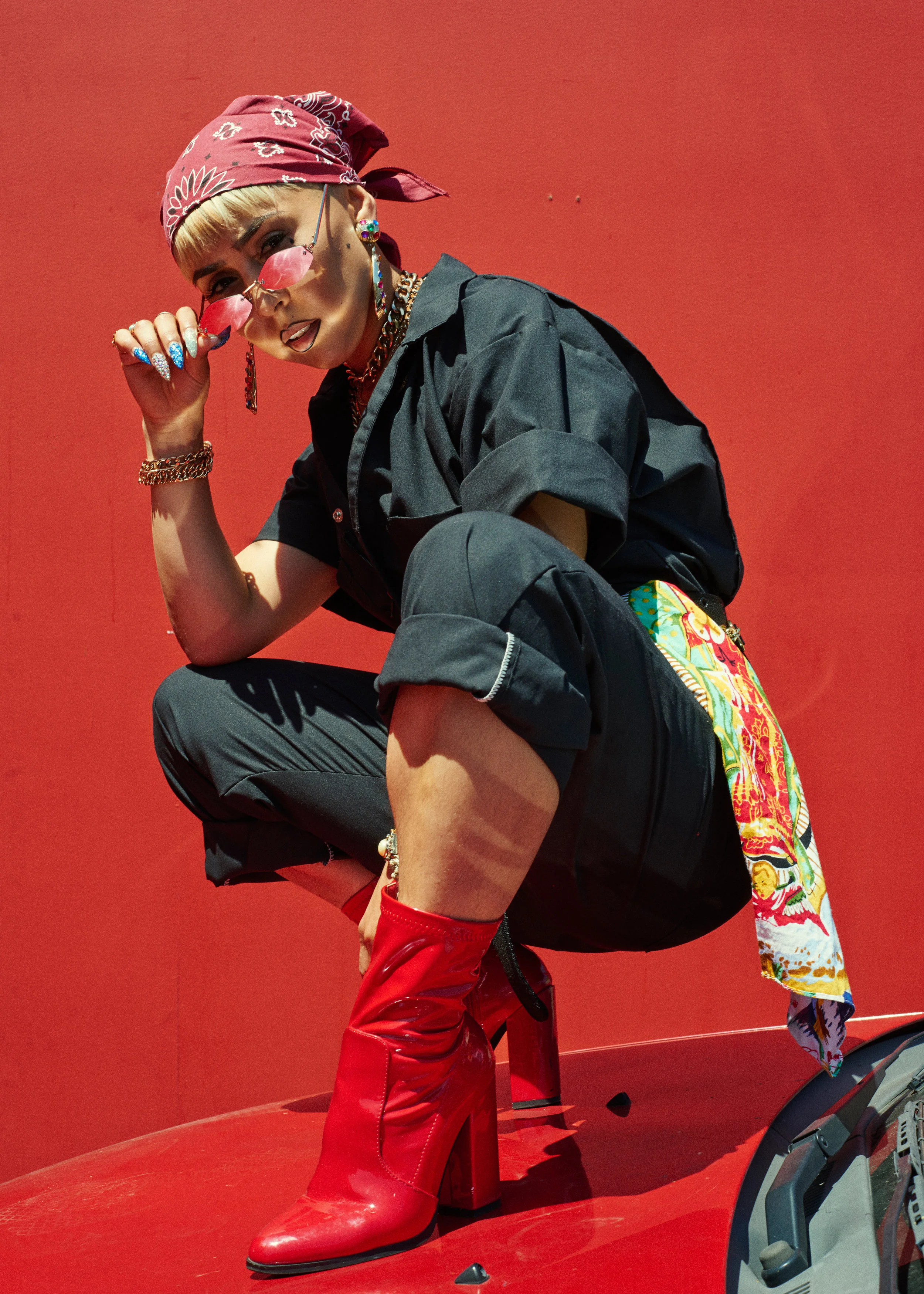 A person squatting on a red surface against a red background, wearing a pink bandana, pink sunglasses, a black shirt, rolled-up black pants, red boots, and colorful accessories.