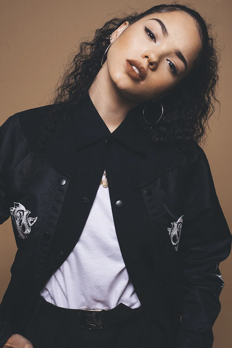 A woman with curly hair wearing a black jacket with animal patches, a white t-shirt, and hoop earrings, posing against a brown background.