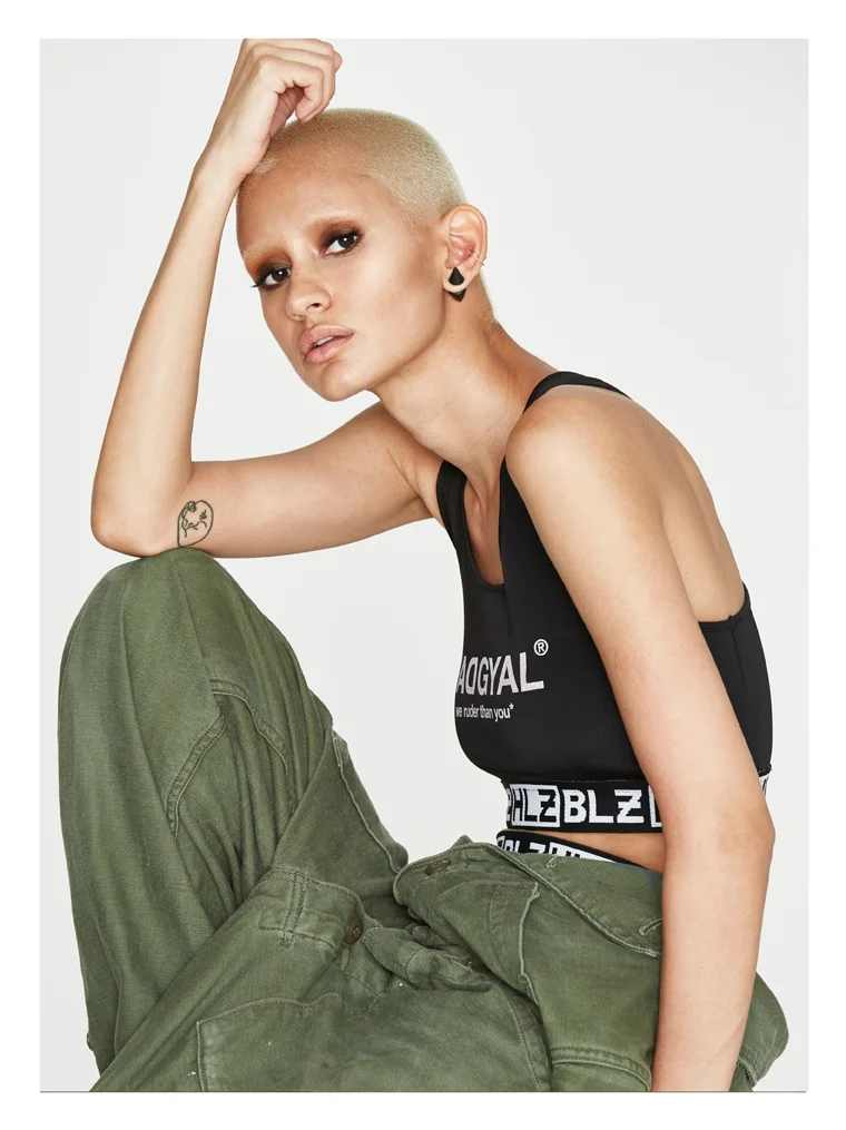 A woman with a shaved head, light skin, and tattoos is sitting, wearing a black sports bra with white text and olive green cargo pants. She has earrings and a visible tattoo on her arm.