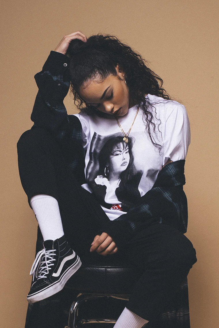 Young woman sitting on a black chair against a beige background, wearing a graphic T-shirt with a woman's face, a plaid jacket, white socks, and Vans sneakers. She has curly hair, a gold necklace, and appears to be deep in thought.