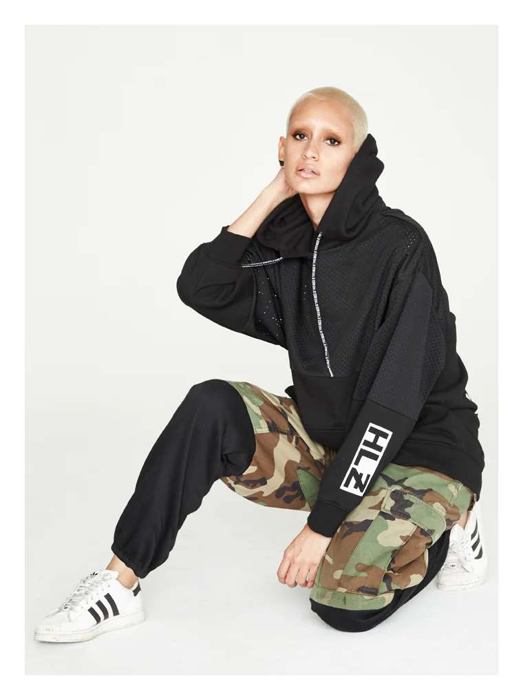 A young woman with bleached blonde hair posing in a black hoodie, camouflage cargo pants, and white sneakers with black stripes.