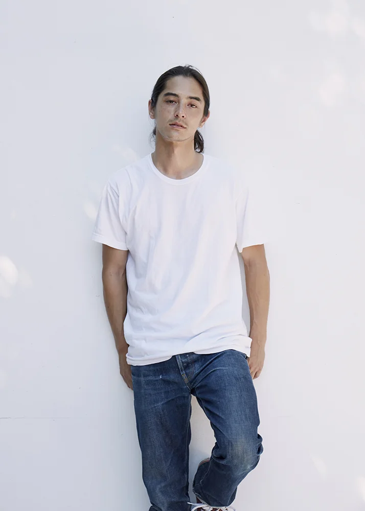 A young man with long dark hair, wearing a white T-shirt and blue jeans, leaning against a white wall with a relaxed expression.