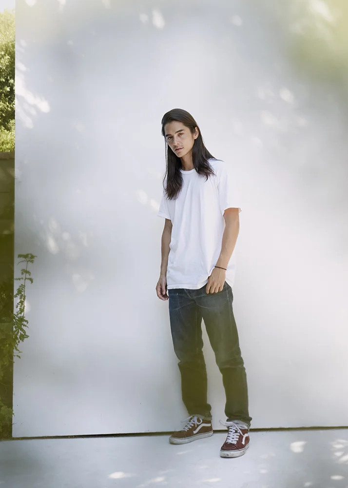 A young man with long dark hair and light skin wearing a white t-shirt, dark jeans, and brown sneakers standing outdoors against a plain white backdrop with shadows from trees. e-commerce, editorial, fashion, vogue, portrait