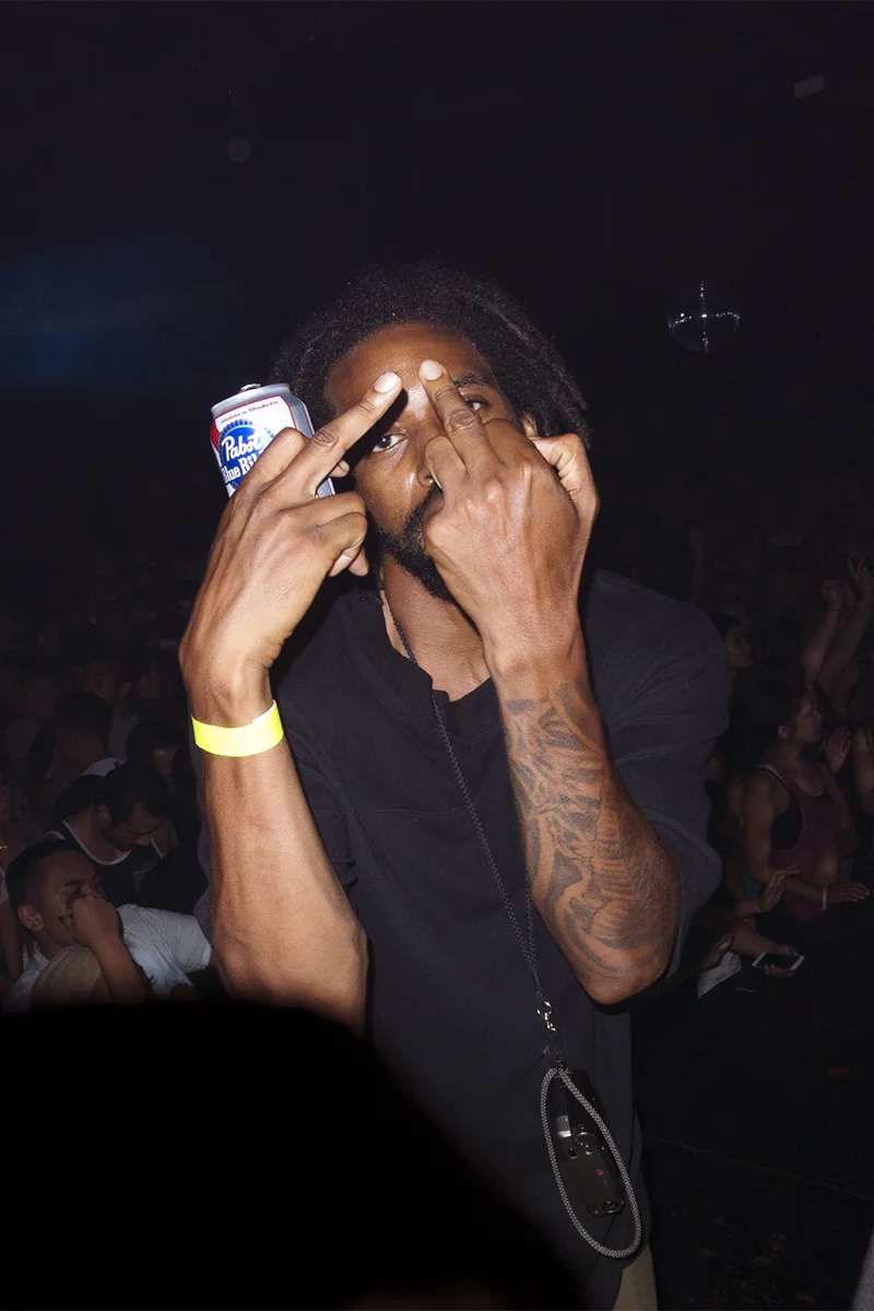 A man with dreadlocks and tattoos making a gesture with his hand while holding a can of Pabst Blue Ribbon beer at a crowded event or concert.