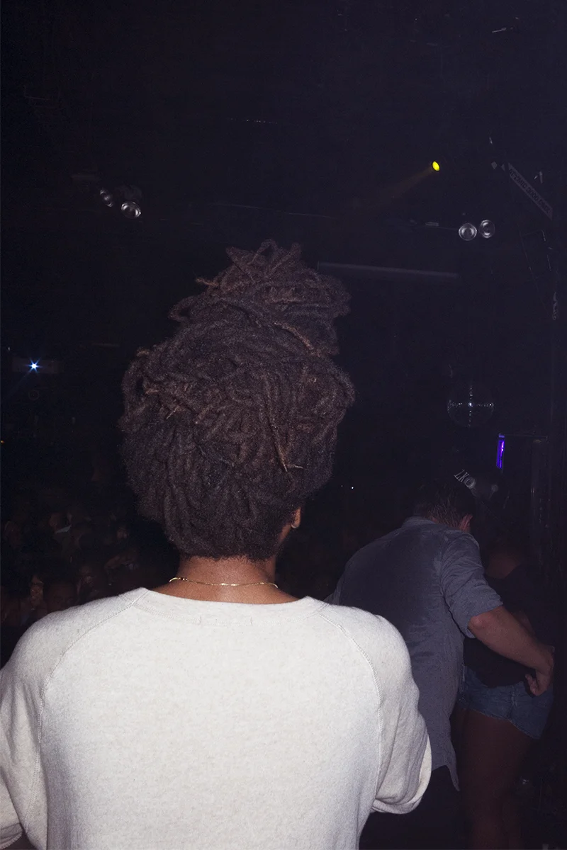 Person with dreadlocks seen from behind in a dark nightclub or concert setting with other people and stage lighting.