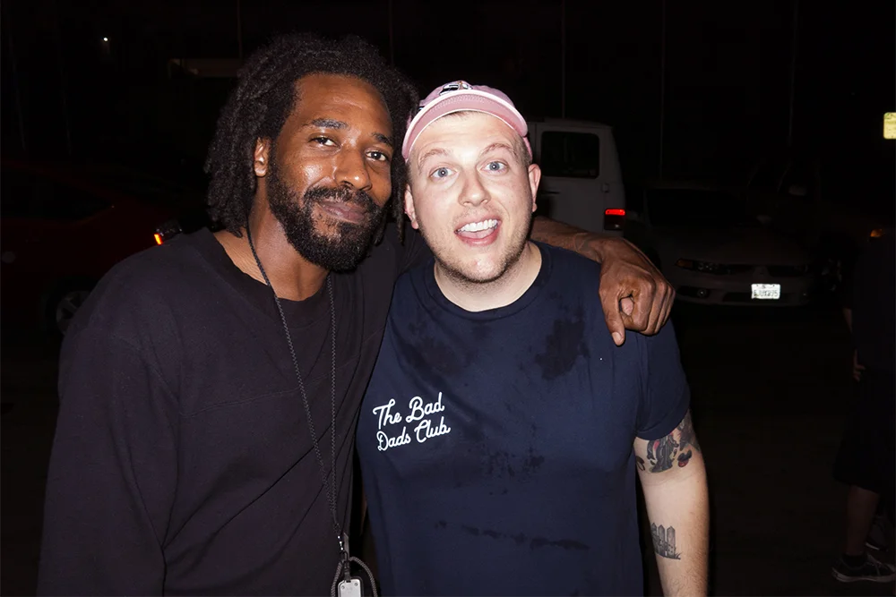 Two men standing together outdoors at night, smiling at the camera, with one man having his arm around the other's shoulder.