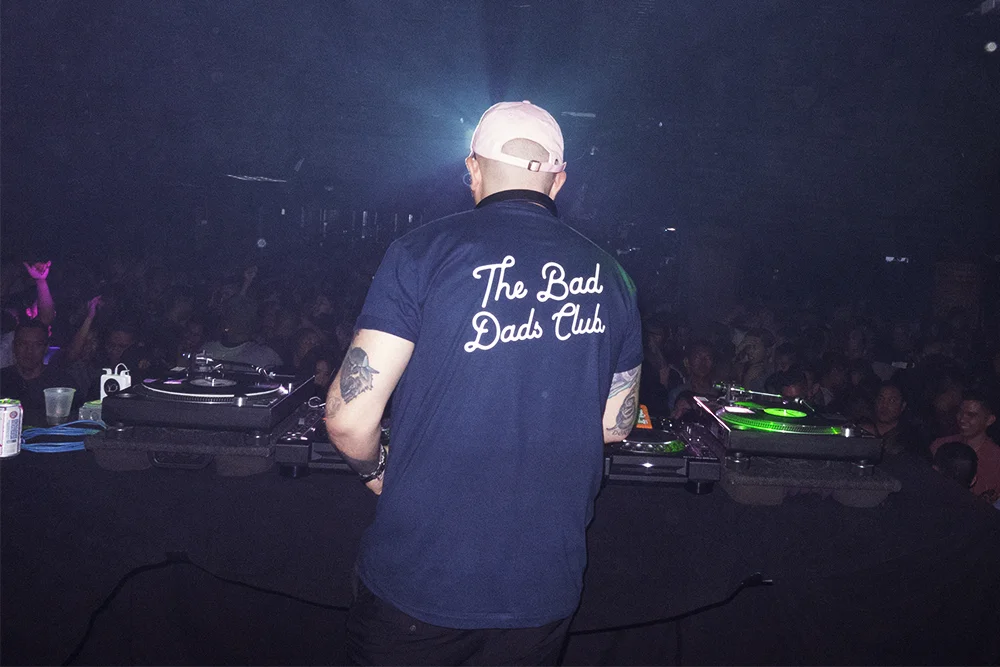 A DJ wearing a navy blue shirt with 'The Bad Dads Club' on the back and a white cap, standing behind DJ equipment at a nighttime event or club with a crowd of people dancing.