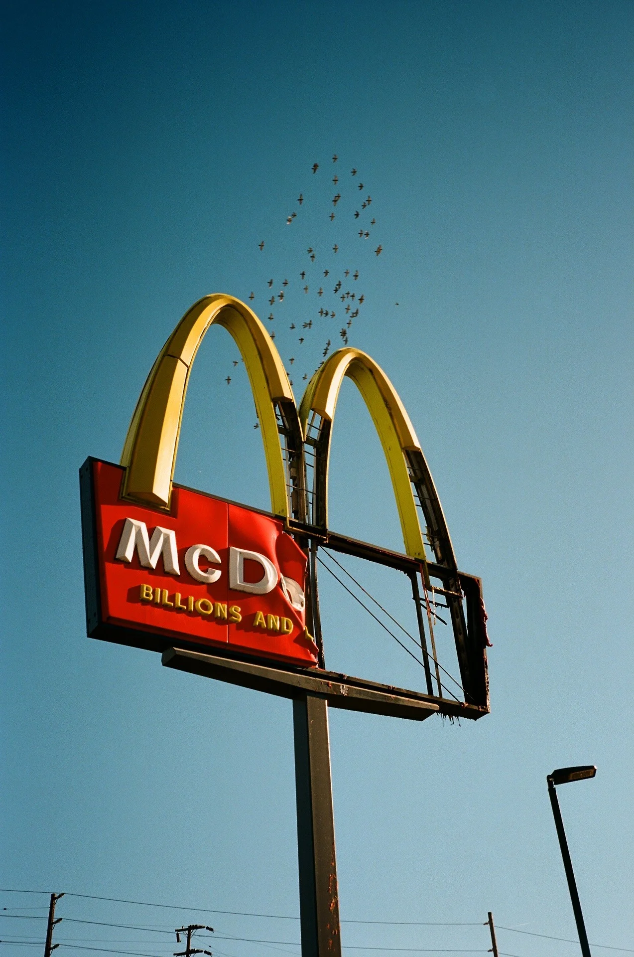 Old, weathered McDonald's sign with missing letters, showing the iconic yellow arches on a blue sky background, with birds flying overhead.