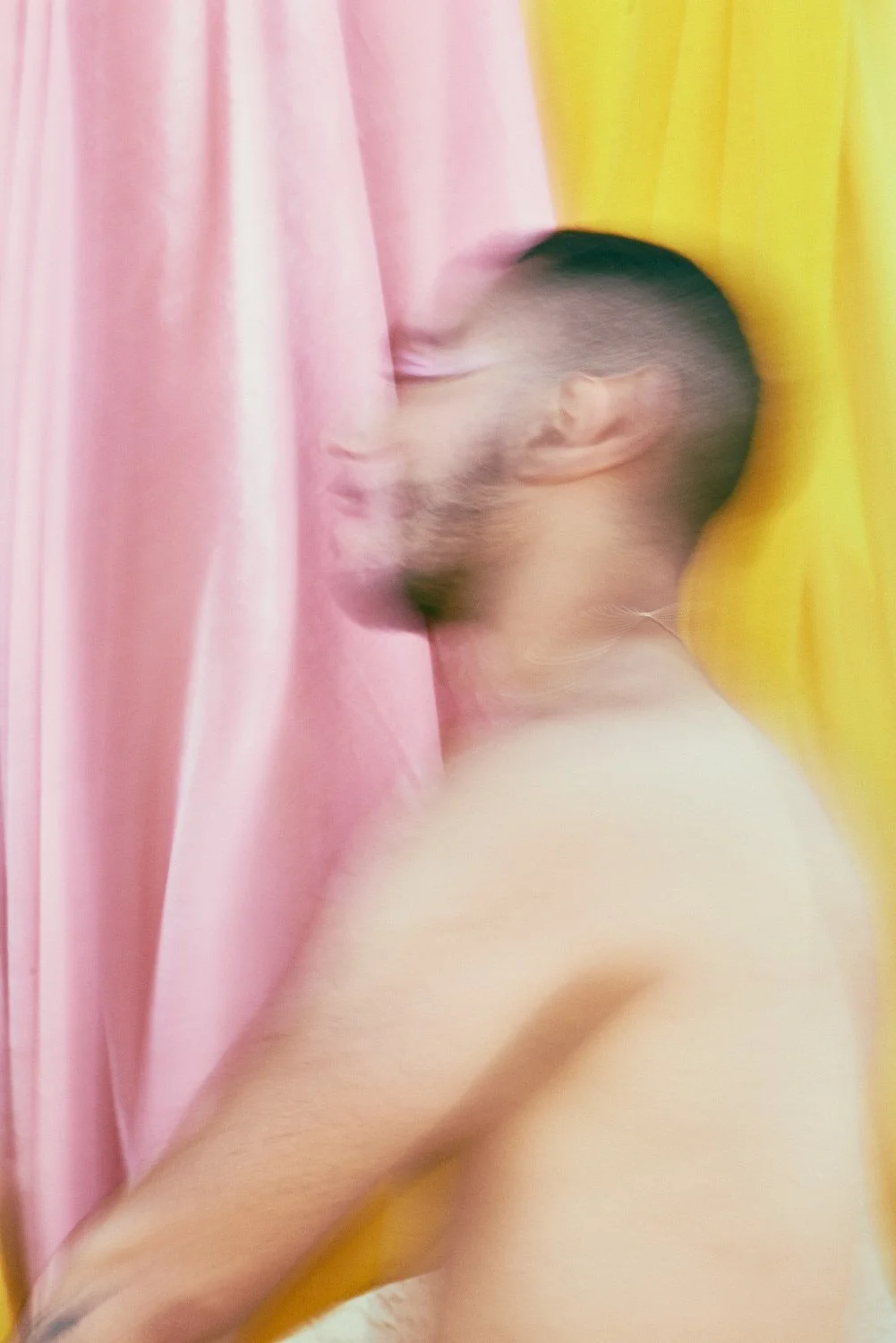 Blurry portrait of a shirtless man with a beard and short hair standing against pink and yellow curtains.