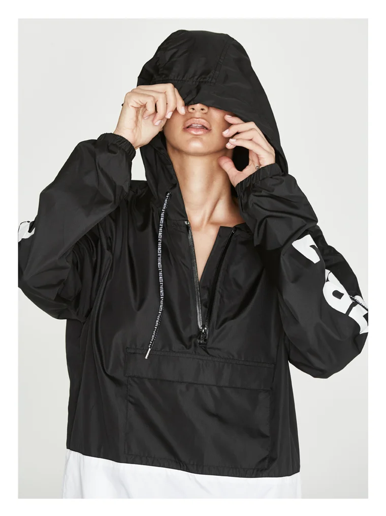 Woman wearing a black hooded windbreaker jacket, partially covering her face, with the hood pulled down over her eyes, and white pants.