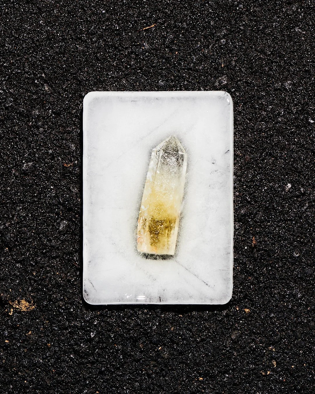A small, clear, quartz crystal encased in a block of ice, placed on a rectangular clear glass plate on a black, textured surface.