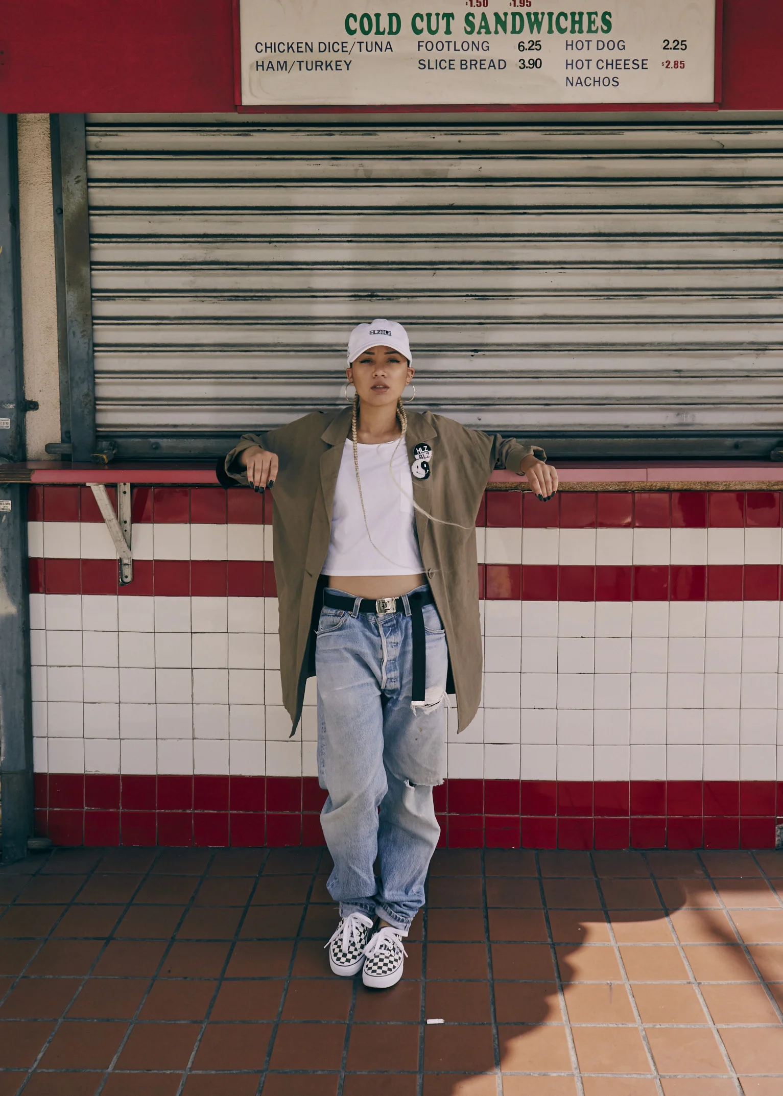 A young woman standing in front of a closed food stand with a red-tile wall, wearing a white baseball cap, an olive green oversized jacket, a white crop top, baggy ripped jeans, and checkerboard Vans sneakers.