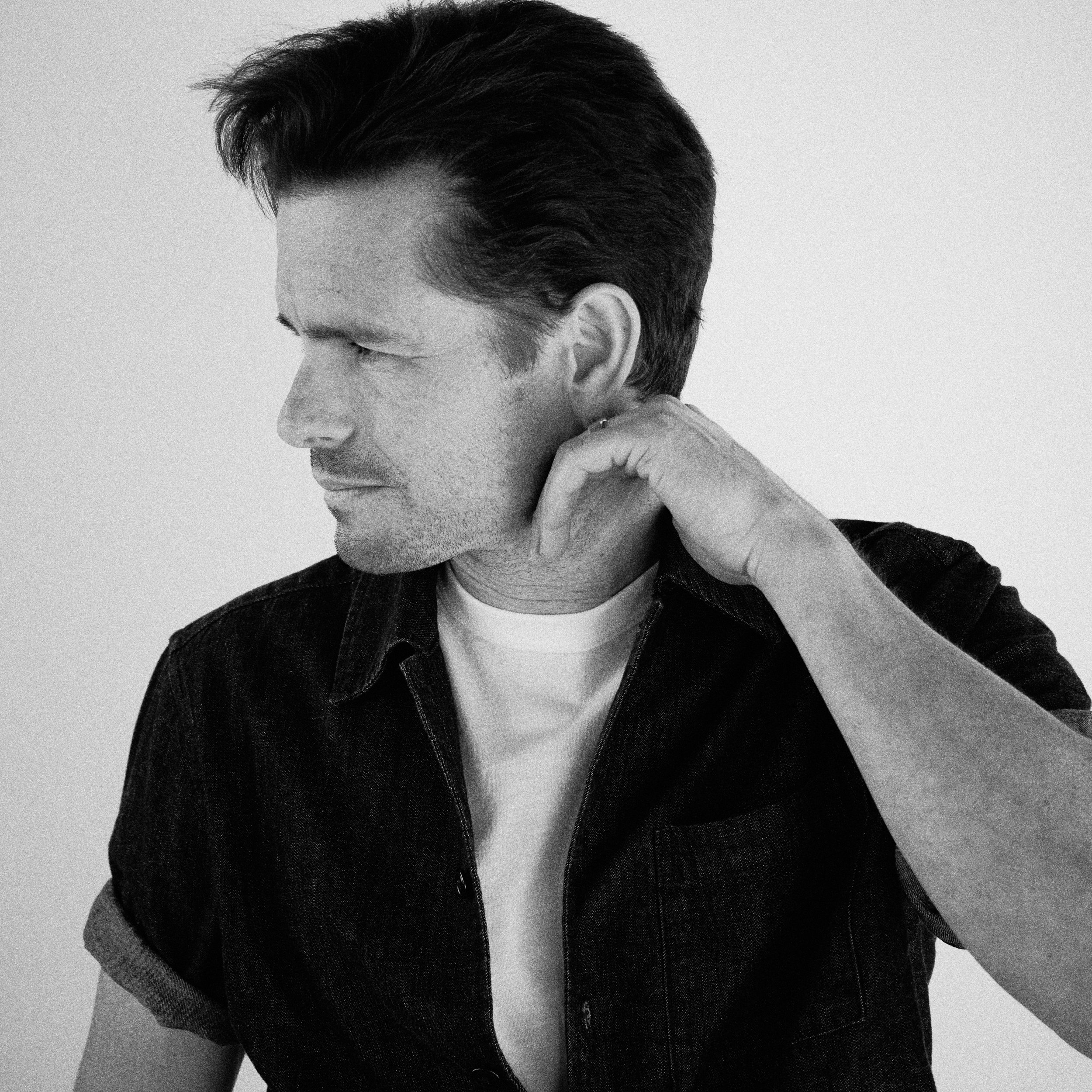 Black-and-white photo of a man with dark hair, wearing a white t-shirt and a dark jacket, with his hand behind his neck, looking to the side