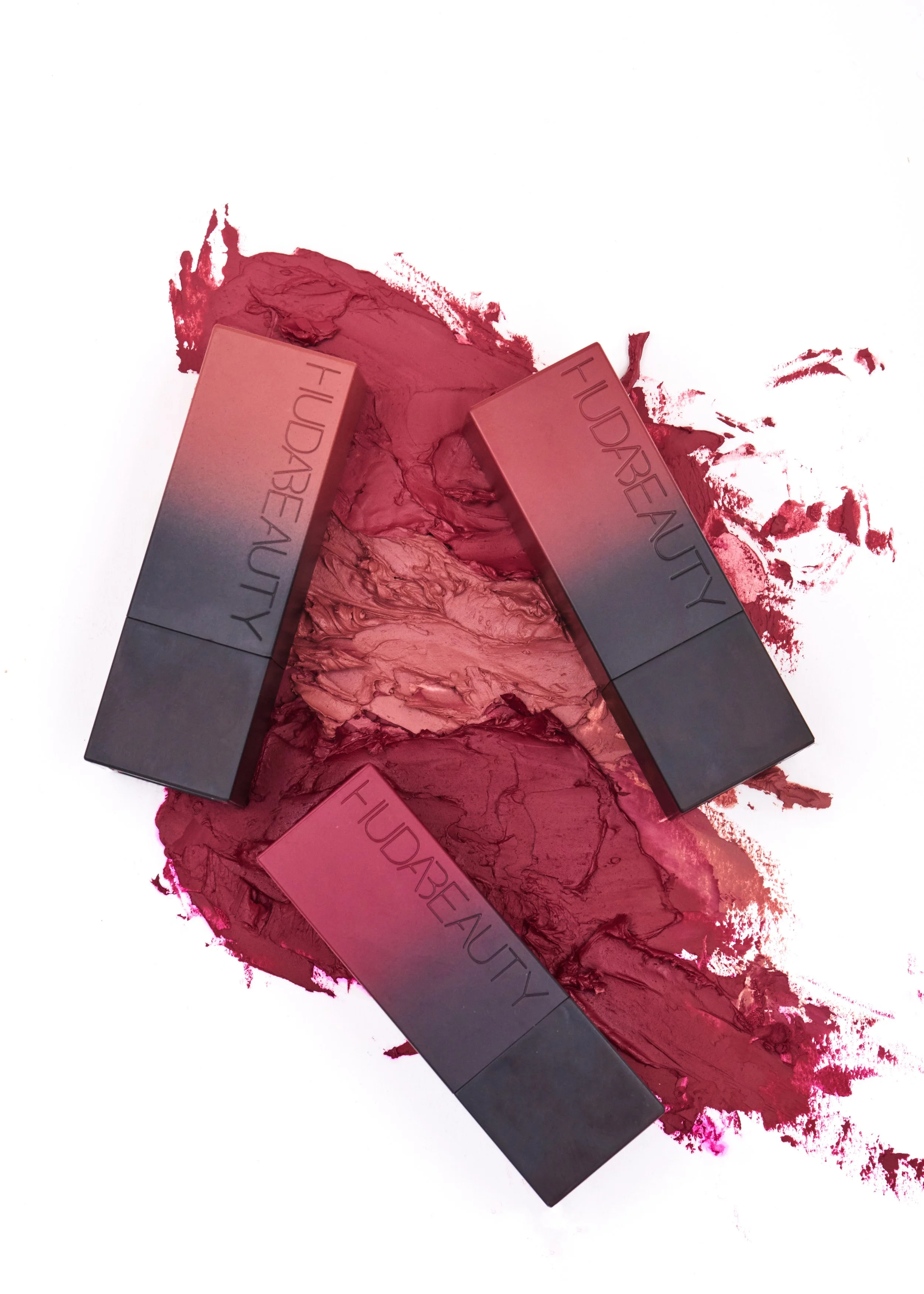 Three Huda Beauty makeup lipsticks lying on a surface with crushed and smeared red and pink lipstick.