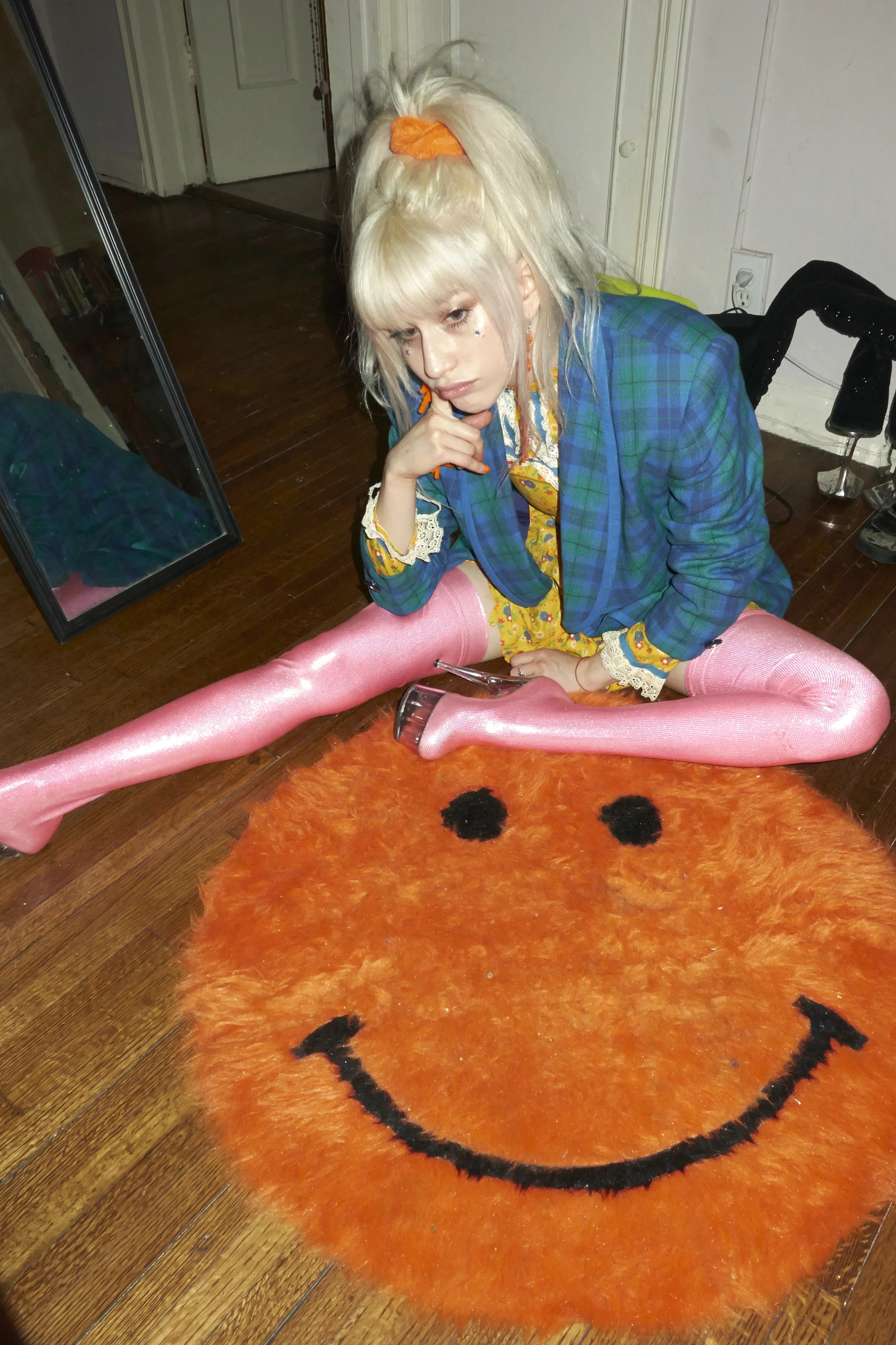 A young woman with platinum blonde hair, wearing a blue plaid blazer, yellow dress, pink shiny over-the-knee stockings, and high heels, is sitting on a round orange smiley face rug on a wooden floor. She has a thoughtful expression, with her hand res