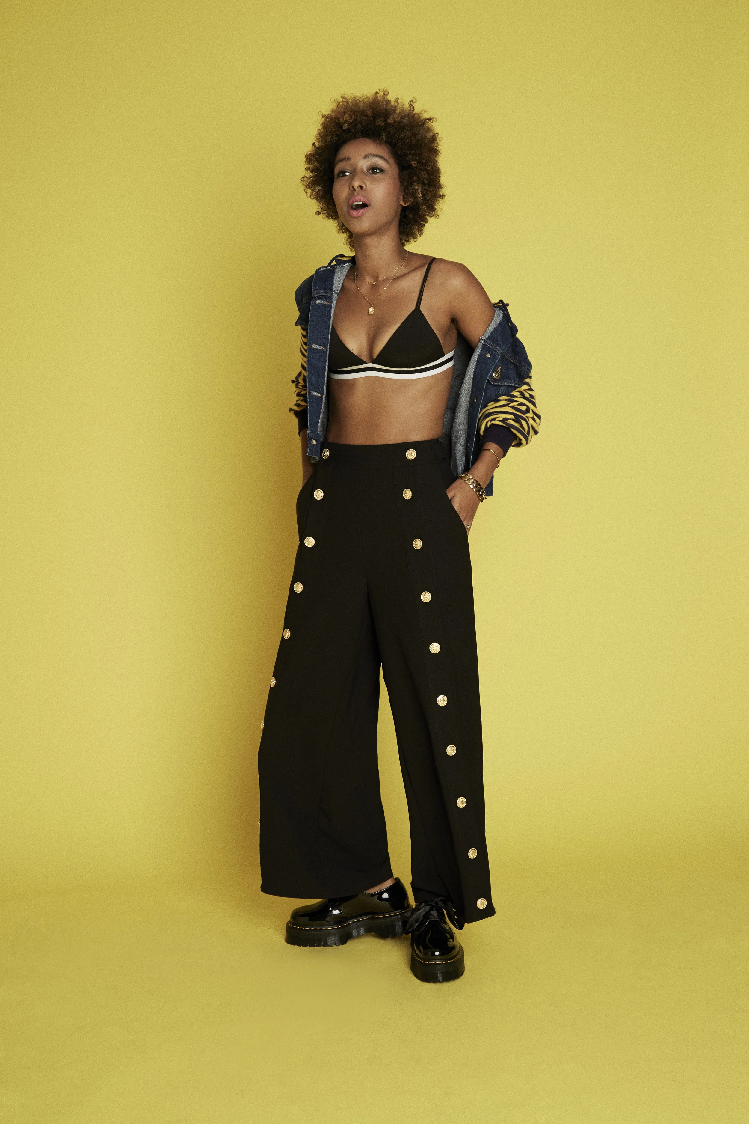 A woman standing against a yellow background, wearing a black bralette with white stripes, high-waisted black pants with gold buttons, black shiny shoes, and a partially unbuttoned denim jacket with a yellow and black patterned inner lining.