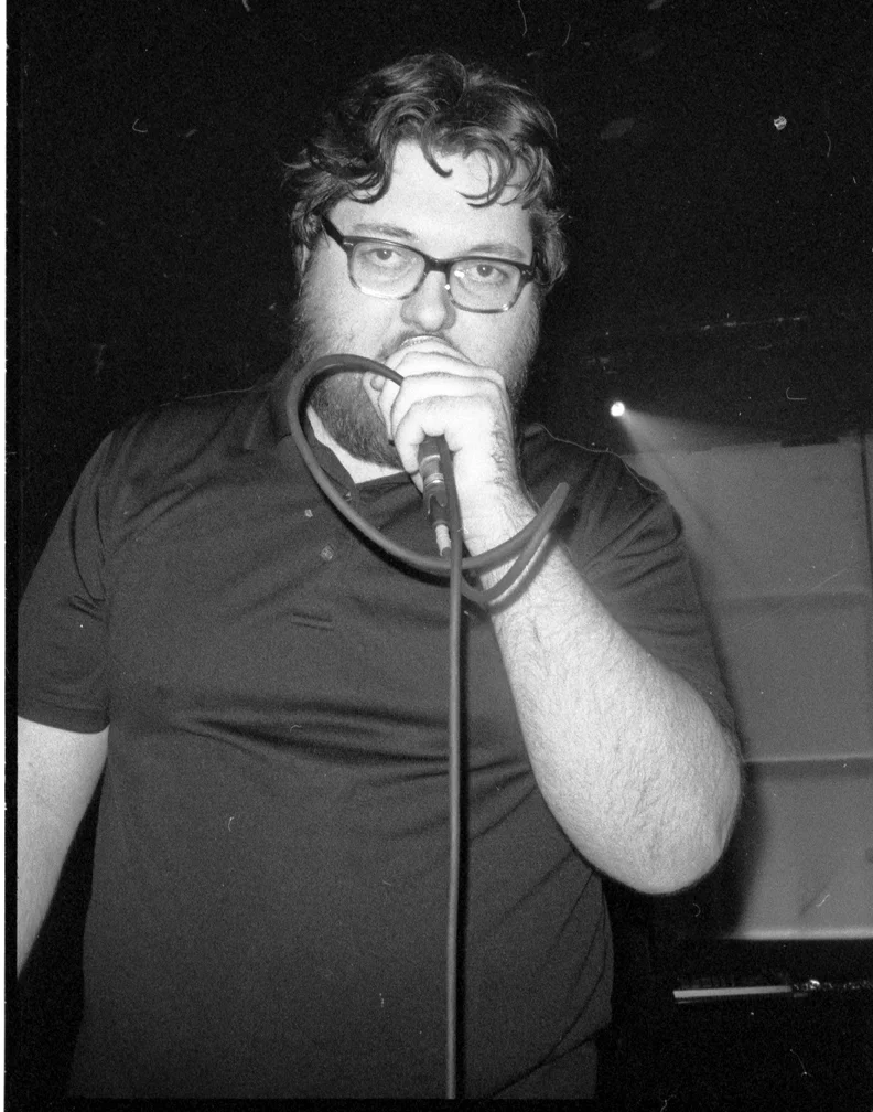 A man with glasses, dark hair, and a beard holding a microphone in front of his mouth, standing in a dark room with a spotlight in the background. music